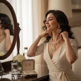 Woman adjusting jewelry in front of a mirror, wearing a beige dress.