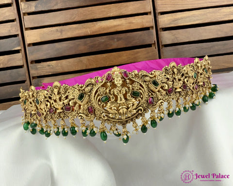 Decorative gold necklace with green and red stones on a white surface with a wooden background