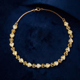 A gold heart-link chain necklace adorned with clear pavé stones, displayed on a textured dark blue velvet background.