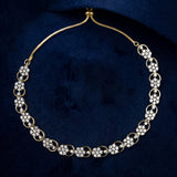 A gold-plated choker necklace with floral and circular links, embellished with clear rhinestones, displayed on a textured dark blue velvet background.