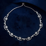 An ornate silver-toned necklace with floral and vine motifs set with clear stones, displayed on a dark navy velvet background.