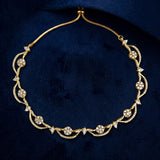 A gold filigree necklace with pavé diamonds, featuring floral and leaf motifs, displayed on a dark navy blue velvet background.