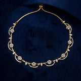 A gold chain necklace with intricate floral and vine motifs set with clear gemstones, displayed on a dark navy blue velvet background.
