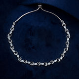 A silver-toned chain necklace with repeating heart-shaped links, each adorned with small clear stones, displayed on a dark navy velvet background.