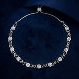 A silver-toned diamond tennis necklace with floral and scroll motifs, displayed on a dark navy blue velvet background.
