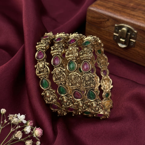 A stack of ornate gold-toned bangles decorated with green and pink teardrop-shaped stones and detailed motifs, arranged on draped burgundy fabric next to a wooden box.
