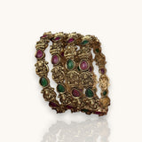 A set of four gold-plated bangles decorated with detailed Ganesha motifs and inlaid green and pink teardrop-shaped stones, displayed on a clean white background with a soft reflection.