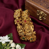 A pair of ornate gold-toned bangles with detailed deity motifs and small red and green stone accents, displayed on draped burgundy fabric next to white flowers and a wooden box.