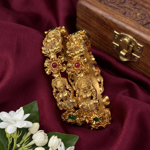 A pair of ornate gold-toned bangles with detailed deity motifs and small red and green stone accents, displayed on draped burgundy fabric next to white flowers and a wooden box.