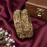 Two ornate gold bangles with floral and bird motifs, decorated with green and pink stones, displayed on a draped burgundy satin fabric next to small white flowers and a wooden box.