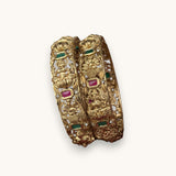 A pair of ornate gold-toned bangles with intricate floral and bird motifs, accented by small green and pink gemstones, displayed on a light cream background.