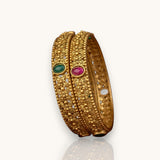 An ornate gold-toned bangle bracelet with intricate floral filigree detailing and two oval-colored stone accents, one green and one pink, displayed on a clean off-white background with a soft reflection below.