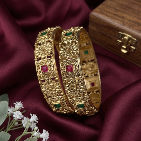 Two ornate gold-toned bangles with floral filigree patterns and red and green stone accents, displayed on draped burgundy fabric next to a wooden box and small white flowers.