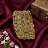 A stack of ornate gold-toned bangles decorated with red and green stone accents, displayed on draped burgundy satin fabric with small pink and white flowers and a wooden box nearby.