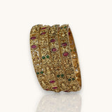 A set of five ornate gold-plated bangles decorated with red and green stones and detailed filigree patterns, displayed on a clean white background with a soft reflection below.