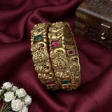 Two ornate gold-toned bangles with carved floral and animal motifs, accented by green and pink gemstones, displayed on draped burgundy satin fabric with white flowers and a wooden box in the background.