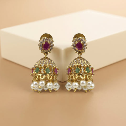 A pair of gold-plated Indian Jhumka earrings with white faux pearls, purple and green stones, and clear crystals, displayed on a beige background with a cream-colored box.