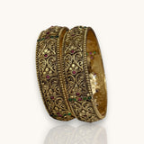 A pair of wide gold-toned bangles with intricate filigree patterns and small red and green stone accents, displayed on a light gray reflective surface.