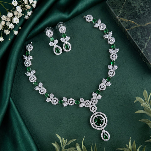 A silver-toned jewelry set featuring a necklace and matching earrings adorned with round and marquise-cut green emeralds, surrounded by clear pavé stones in floral motifs, arranged on a dark green satin fabric background with decorative white flowers.