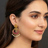 A close-up of a woman wearing a traditional Indian gold jhumka earring with red and green enamel, pearl dangling bells, and a green stone accent.