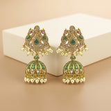 A pair of gold-plated Indian jhumka earrings with green enamel, clear rhinestones, and small pink accent stones, displayed on a beige background with a white block.