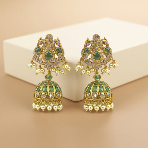 A pair of gold-plated Indian jhumka earrings with green enamel, clear rhinestones, and small pink accent stones, displayed on a beige background with a white block.