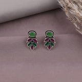 A pair of silver-toned drop earrings with oval green stones at the top and teardrop-shaped green and purple stones below, displayed on a mauve surface with dried grass in the corner.