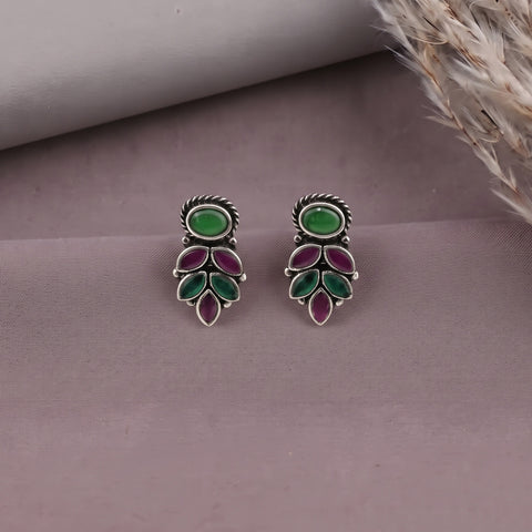 A pair of silver-toned drop earrings with oval green stones at the top and teardrop-shaped green and purple stones below, displayed on a mauve surface with dried grass in the corner.