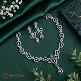 Silver-toned jewelry set with a necklace and matching earrings featuring intricate filigree patterns and green stone accents, arranged on a dark green satin fabric with small white flowers and green leaves.