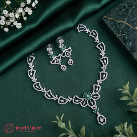 Silver-toned jewelry set with a necklace and matching earrings featuring intricate filigree patterns and green stone accents, arranged on a dark green satin fabric with small white flowers and green leaves.