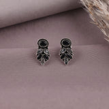 A pair of silver-toned dangle earrings with black and clear stones, displayed on a dusty mauve fabric background with dried grass as a prop.