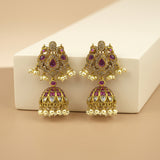 A pair of ornate gold-toned Indian jhumka earrings with purple stones and clear crystal accents, displayed on a white rectangular block against a beige background.