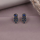A pair of silver-toned dangle earrings with oval and marquise-cut deep blue stones, displayed on a textured mauve surface with dried grass in the corner.