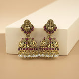 A pair of ornate Indian jhumka earrings with antique gold-plated metalwork, set with deep red gemstones and dangling white pearl beads, displayed on a white rectangular block against a beige background.