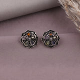 A pair of silver-toned floral stud earrings with multicolored gemstone accents, displayed on a textured mauve surface with a neutral fabric roll and dried grass in the background.