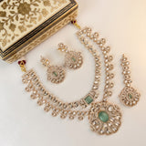 An ornate Indian ethnic jewelry set featuring a necklace, earrings, and a matching box. The jewelry is made of silver-toned metal with clear stones and green stone accents, displayed on a light beige background next to a decorative cream and gold box.