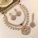 An ornate Indian jewelry set featuring a silver-tone necklace, matching earrings, and a dangling earring, decorated with clear stones and pink stone accents, displayed on a cream-colored background next to a decorative book.
