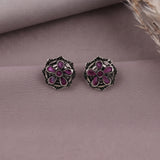 A pair of silver-toned floral stud earrings with purple enamel petals, displayed on a textured mauve surface with a dried feather in the corner.
