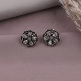A pair of silver-toned floral stud earrings with clear teardrop-shaped stones, displayed on a dusty mauve surface with dried grass in the corner.