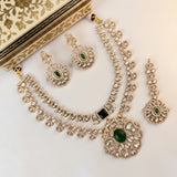 An ornate Indian jewelry set featuring a silver-tone necklace, matching earrings, and a bracelet, embellished with clear crystals and green stones, displayed on a cream-colored patterned box.