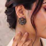 A close-up of a woman wearing a large, ornate silver-toned flower-shaped earring with a small amber accent, shown on her earlobe.