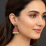 A close-up of a woman's ear wearing a gold-toned traditional Indian jhumka earring embellished with pink stones, green stones, and small white pearl-like beads.