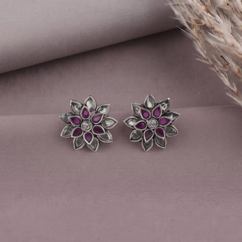 A pair of silver-toned flower-shaped stud earrings with purple gemstone petals and a clear central stone, displayed on a dusty mauve fabric background with dried grass in the corner.