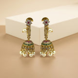 A pair of ornate gold-toned Indian jhumka earrings with white faux pearls, green and red stones, and crystal accents, displayed on a beige background with a white block.