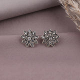 A pair of silver-toned flower-shaped stud earrings with clear teardrop stones, displayed on a dusty mauve fabric background with dried grass in the corner.
