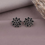 A pair of silver-toned lotus flower stud earrings with dark inlay details, displayed on a textured mauve surface with dried grass in the corner.