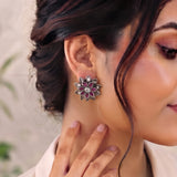A close-up of a woman wearing a silver-toned flower-shaped stud earring with pink gemstone accents, with her hand resting near her neck.