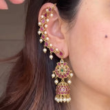 A close-up of a person's ear wearing an ornate gold-toned jhumka-ear cuff earring decorated with pinkish-red stones, white pearls, and a small green stone accent.