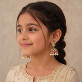 A young girl with dark hair in a braid wearing ornate gold jhumka earrings with pink and green stone accents and white pearl drops, paired with a cream embroidered traditional outfit against a neutral beige background.