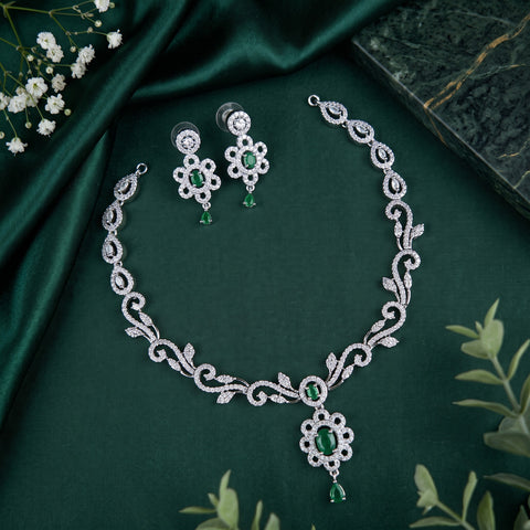 Silver-toned jewelry set with a necklace and matching earrings, featuring silver floral and scroll motifs set with clear stones and green emerald-cut accents, displayed on a dark green satin fabric.